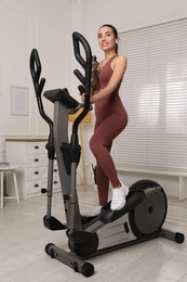 Photo of Happy young woman with bottle of water on elliptical machine at home