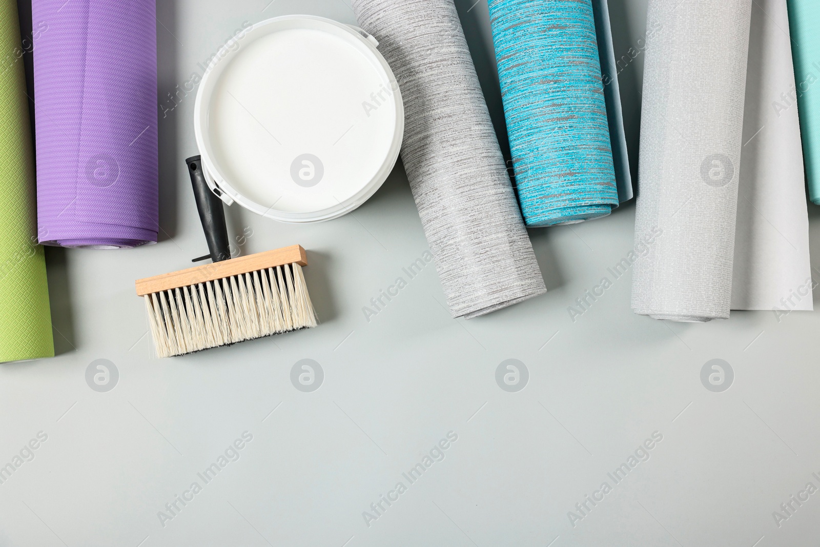 Photo of Different wallpaper rolls, brush and bucket with glue on light grey background, flat lay. Space for text