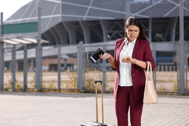 Being late. Worried businesswoman with suitcase and passport looking at her watch outdoors, space for text