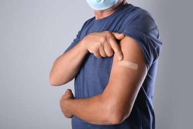 Senior man in protective mask pointing at arm with bandage after vaccination on grey background, closeup