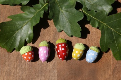 Photo of Colorful painted acorns with polka dot pattern and green oak leaves on wooden table, flat lay