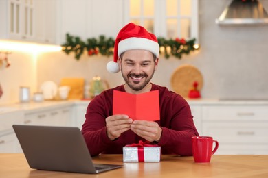 Photo of Celebrating Christmas online with exchanged by mail presents. Happy man reading greeting card during video call on laptop at home