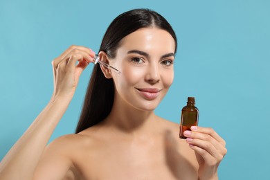 Beautiful young woman applying serum onto her face on light blue background