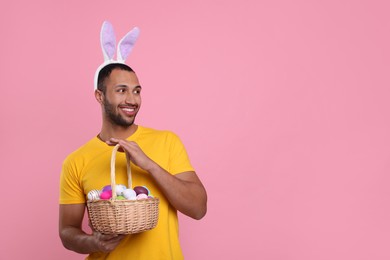 Photo of Happy African American man in bunny ears headband holding wicker basket with Easter eggs on pink background. Space for text