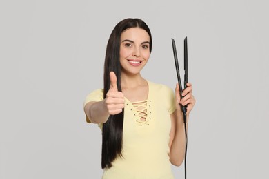 Photo of Beautiful happy woman with hair iron showing thumbs up on light grey background