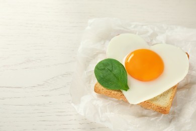 Heart shaped fried egg with toast and spinach on white wooden table. Space for text