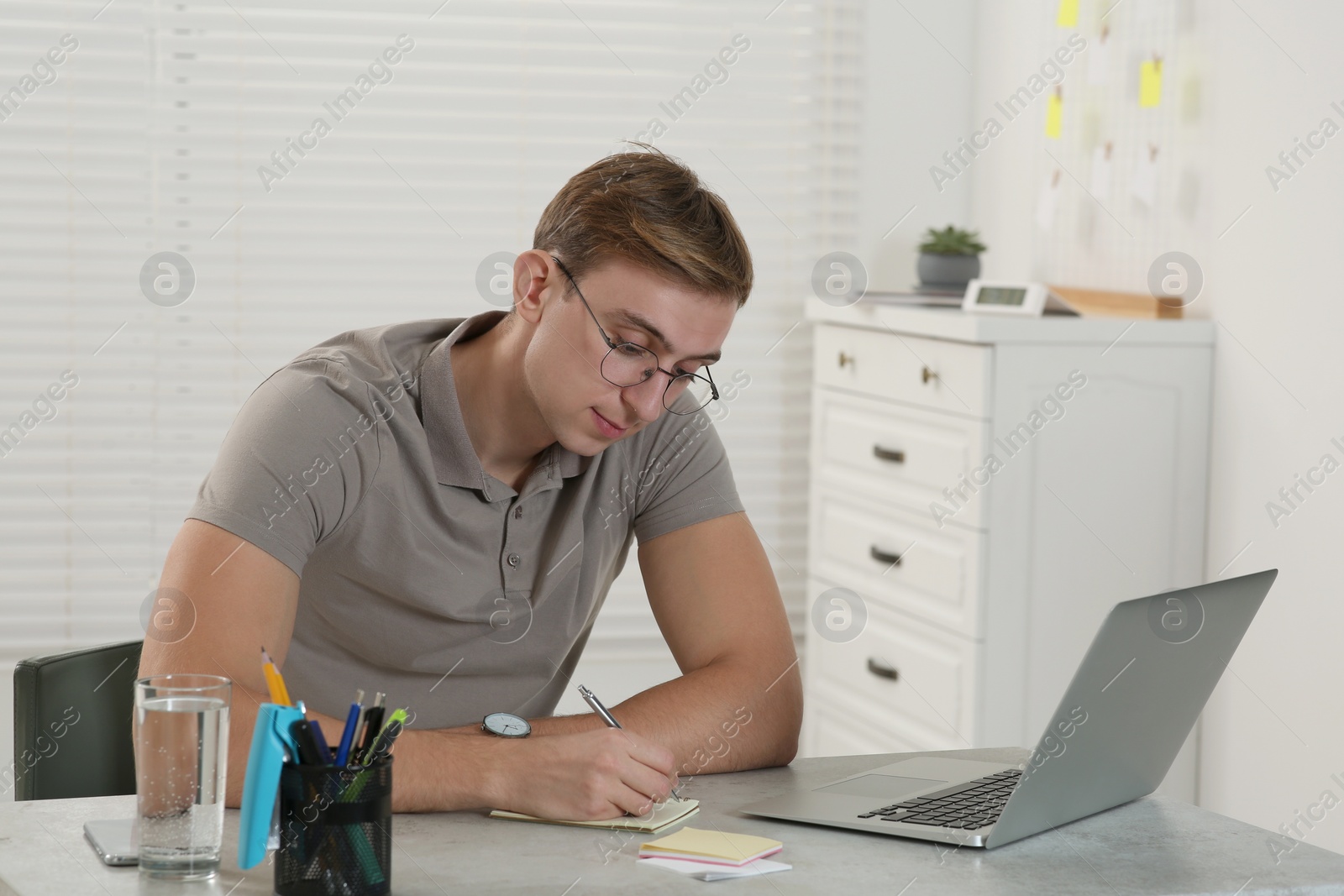 Photo of Young man with laptop writing in notebook at table indoors