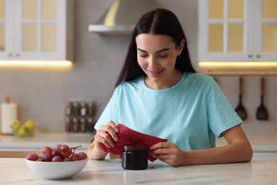Photo of Woman packing jar of jam into beeswax food wrap at table in kitchen