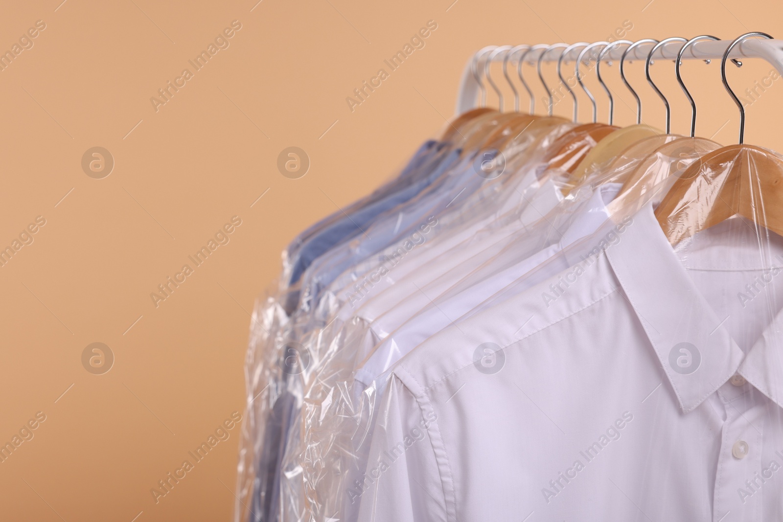 Photo of Dry-cleaning service. Many different clothes in plastic bags hanging on rack against beige background, closeup and space for text
