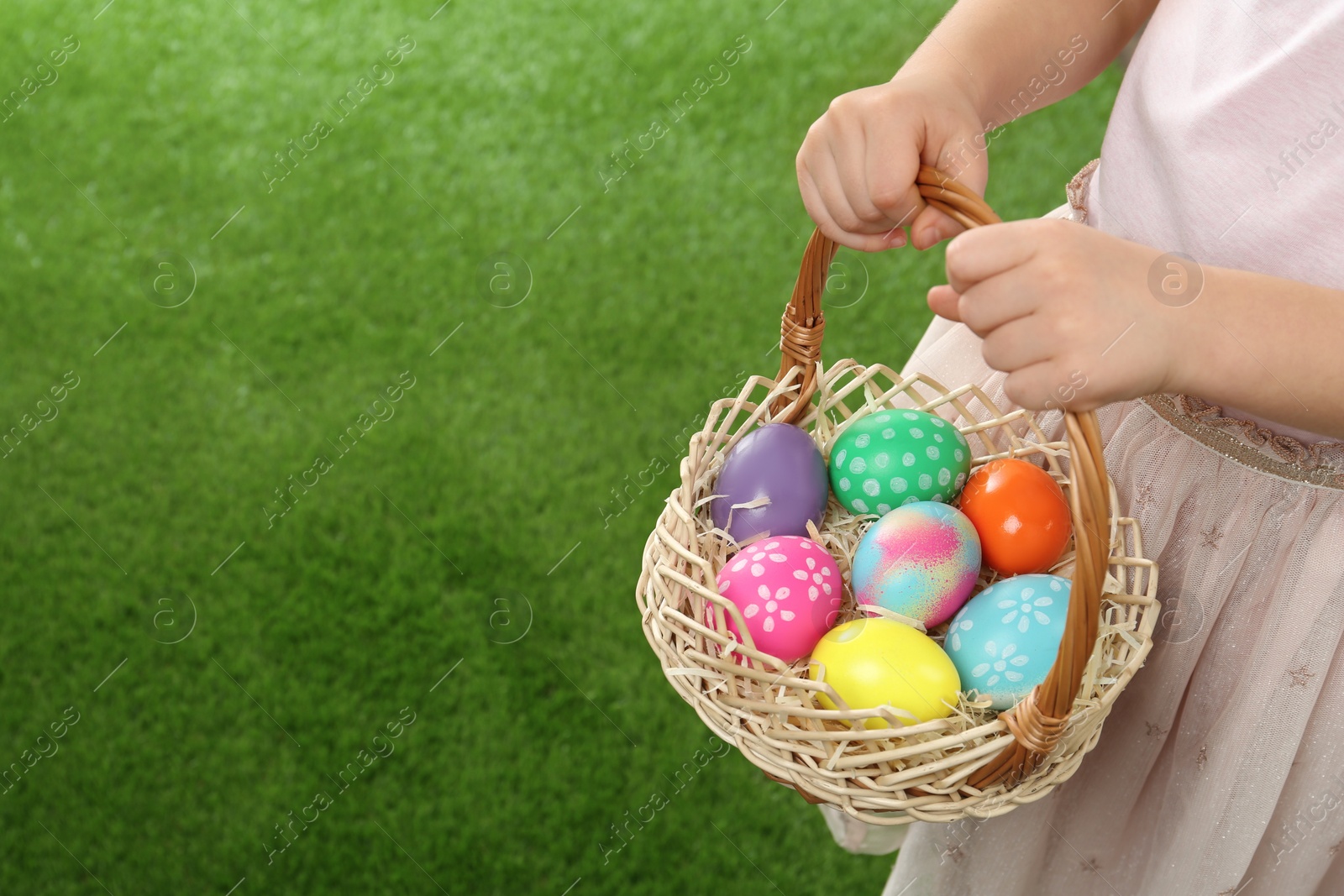 Photo of Little girl with basket full of Easter eggs on green grass, closeup. Space for text