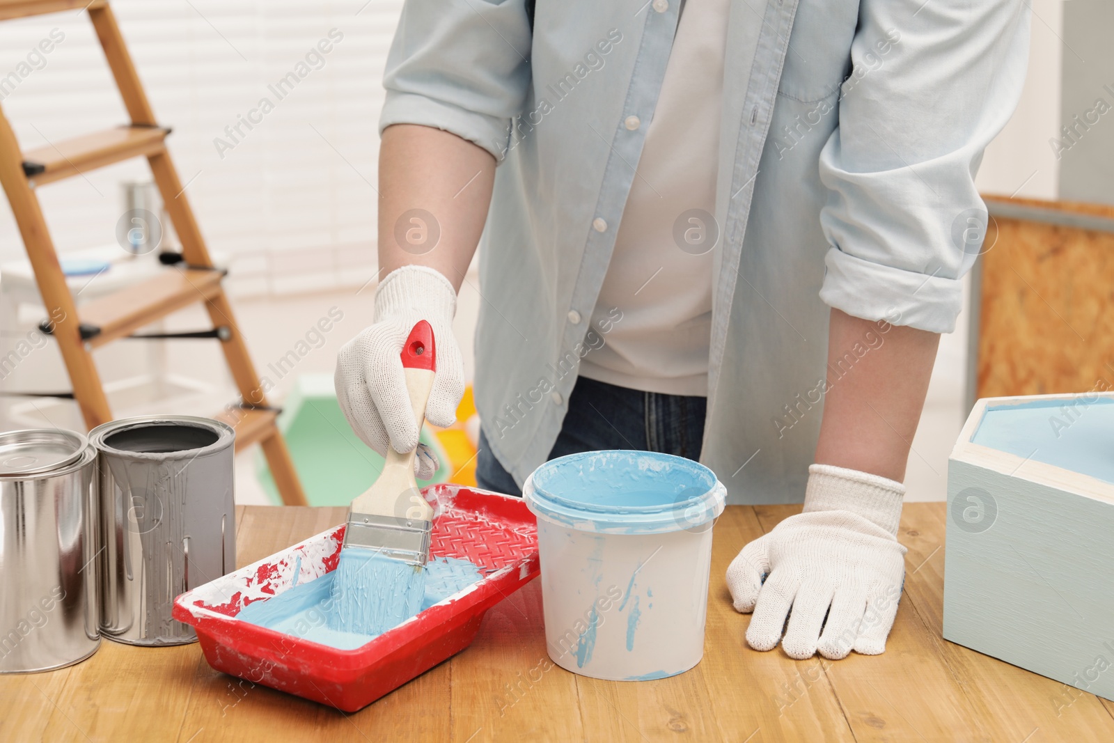 Photo of Man taking light blue paint with brush from tray at wooden table indoors, closeup