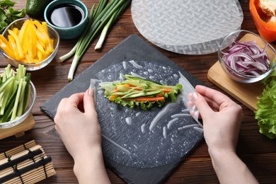 Photo of Woman making spring roll at wooden table with products, closeup