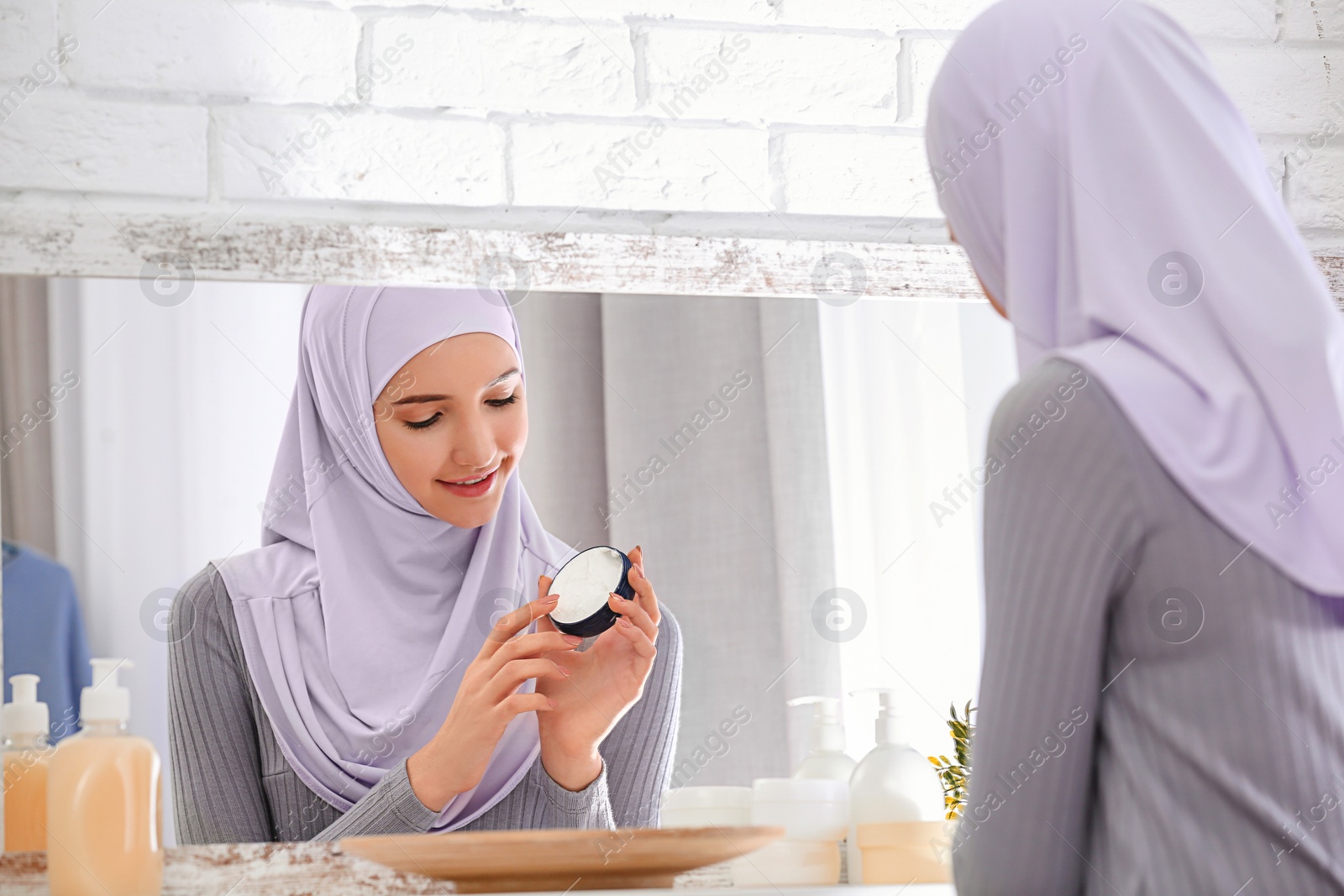 Photo of Beautiful Muslim girl with acne problem using cream while looking in mirror indoors