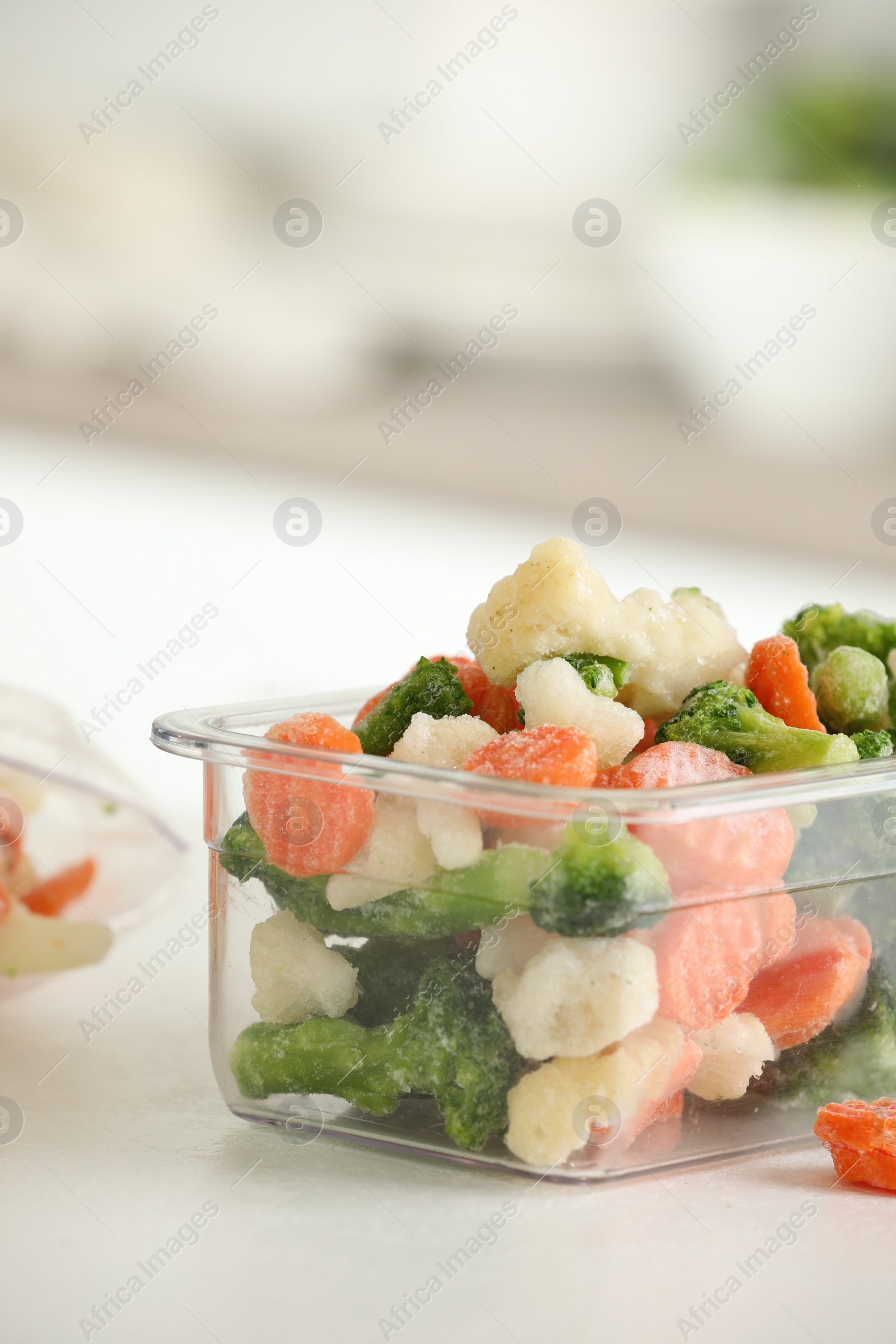 Photo of Mix of different frozen vegetables in plastic container on white textured countertop in kitchen, closeup. Space for text