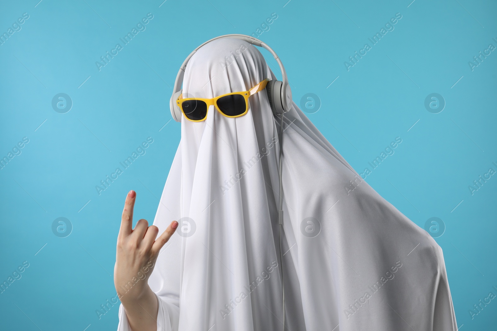 Photo of Stylish ghost. Person covered with white sheet in sunglasses and headphones showing rock sign on light blue background