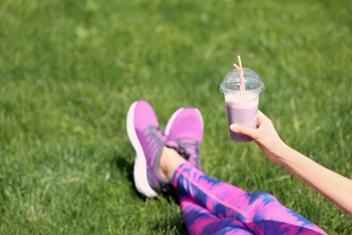 Young woman in sportswear with plastic cup of healthy smoothie on grass outdoors