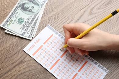 Woman filling out lottery ticket with pencil and money on wooden table, closeup