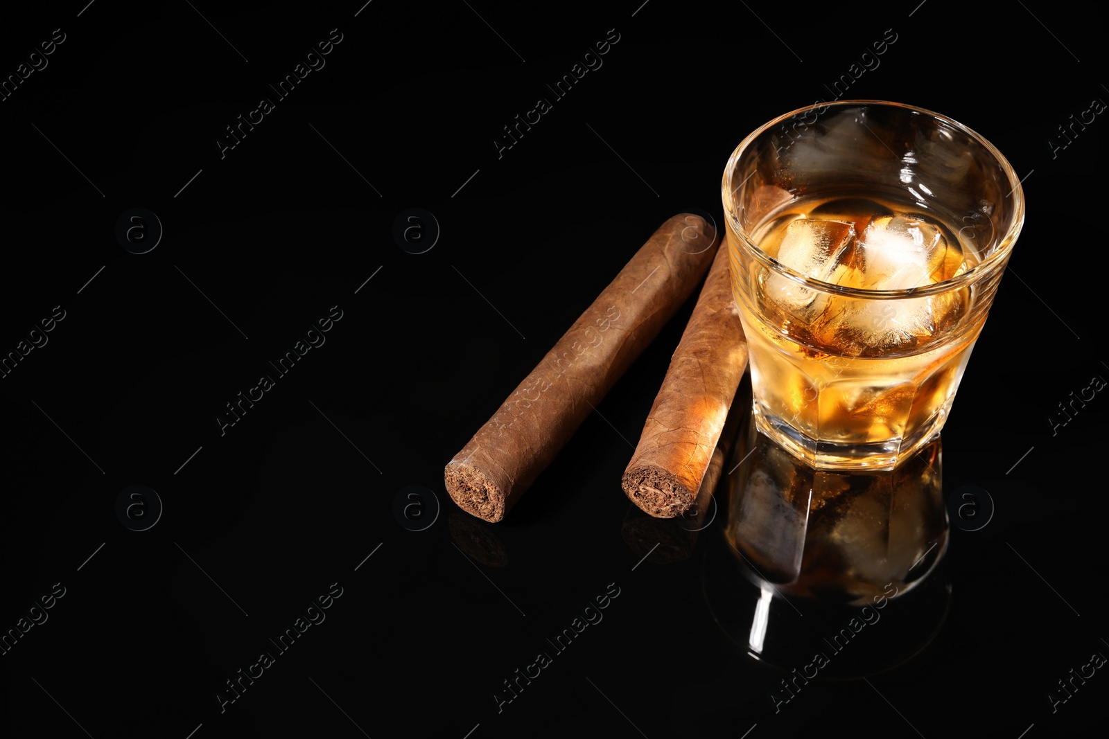 Photo of Glass of whiskey with ice cubes and cigars on black mirror surface. Space for text