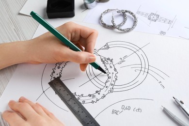 Photo of Jeweler drawing sketch of elegant earrings on paper at white wooden table, closeup