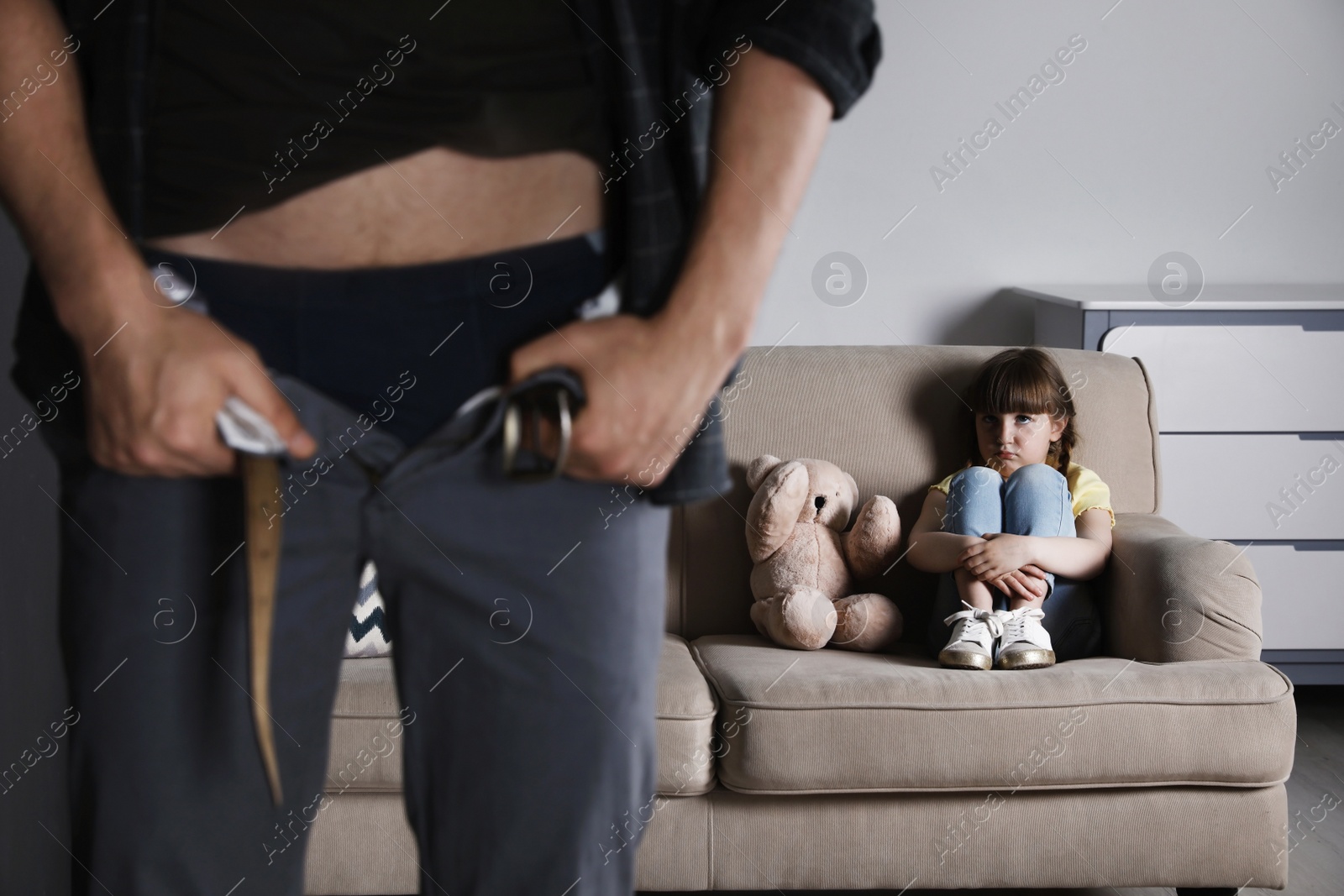 Photo of Man with unzipped pants standing near scared little girl on sofa indoors. Child in danger