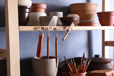 Photo of Set of different crafting tools and clay dishes on wooden rack in workshop, closeup