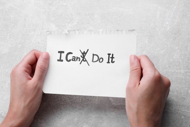 Photo of Motivation concept. Woman holding paper with changed phrase from I Can't Do It into I Can Do It by crossing over letter T at grey table, closeup