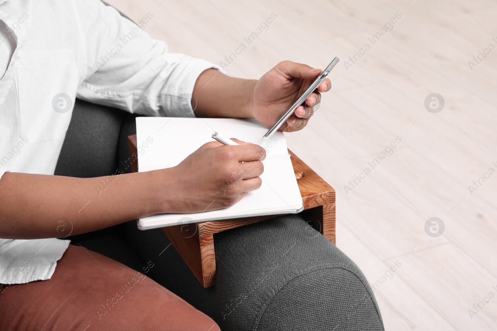 Photo of Man using smartphone while writing in notebook on sofa armrest wooden table at home, closeup