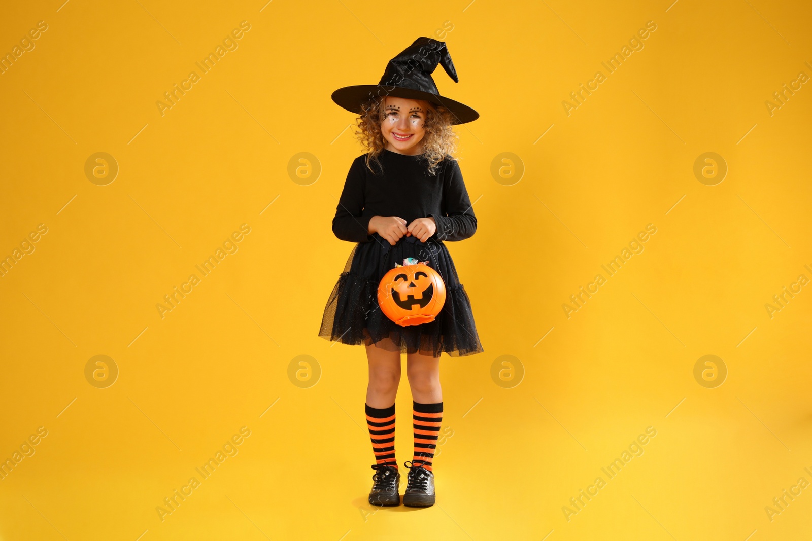 Photo of Cute little girl with pumpkin candy bucket wearing Halloween costume on yellow background