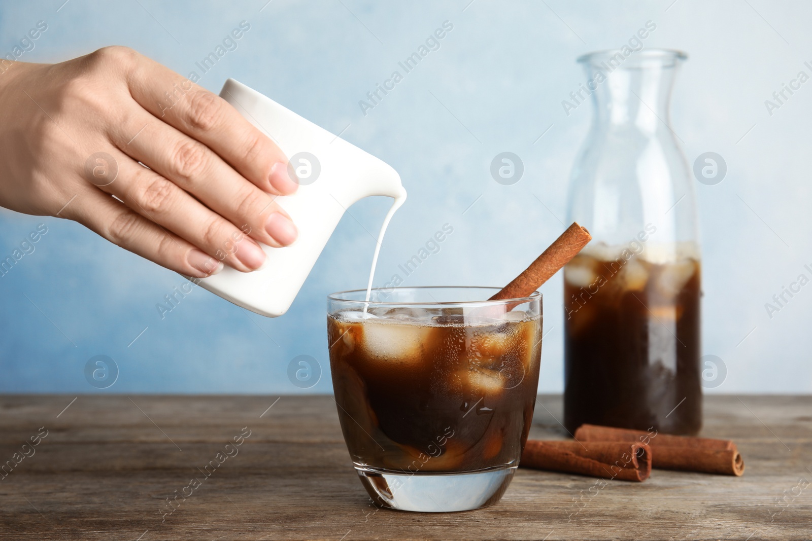 Photo of Woman pouring cream into glass of coffee with ice cubes on table against color background