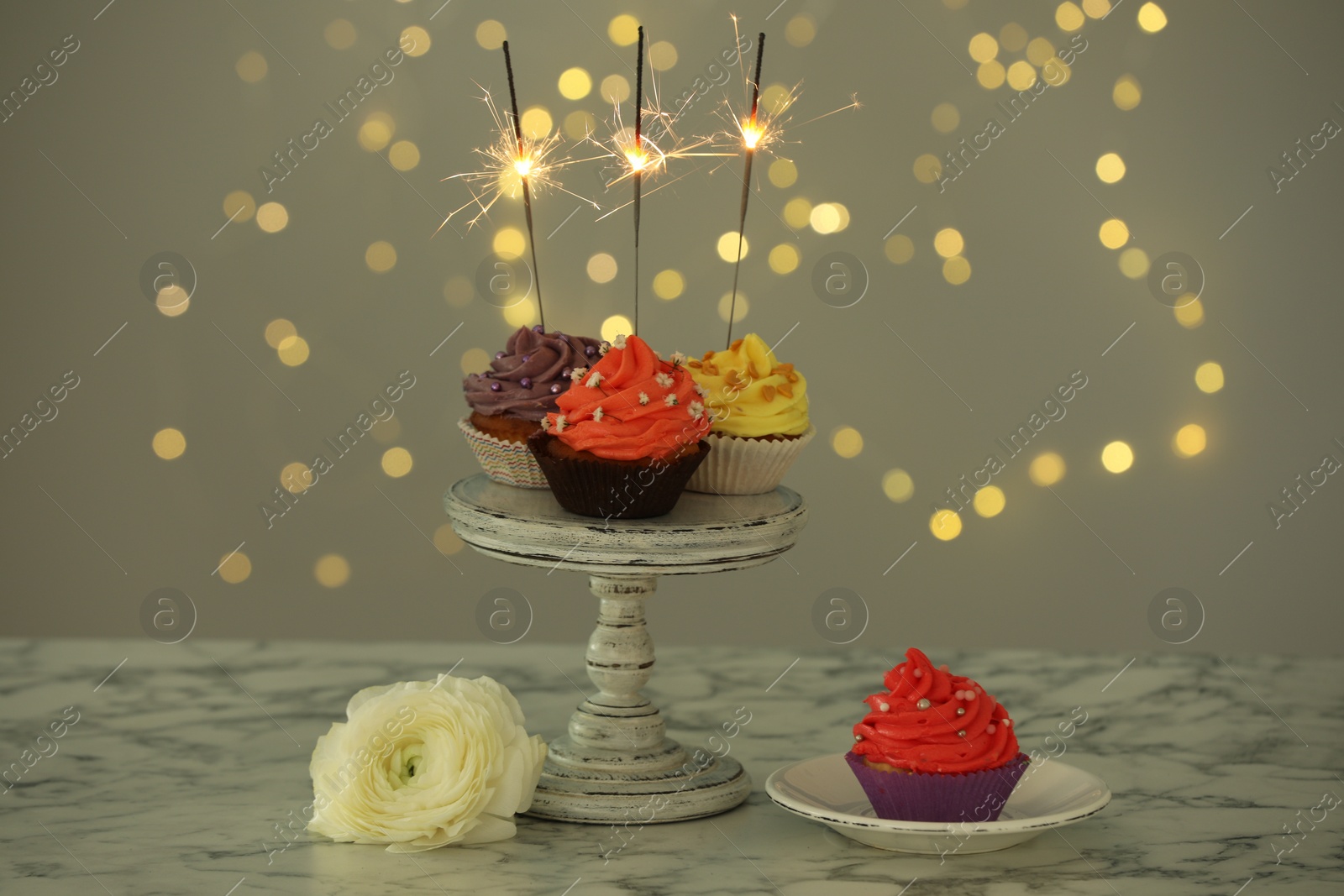 Photo of Different colorful cupcakes with sparklers on white marble table against blurred lights