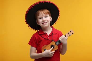Photo of Cute boy in Mexican sombrero hat playing ukulele on yellow background