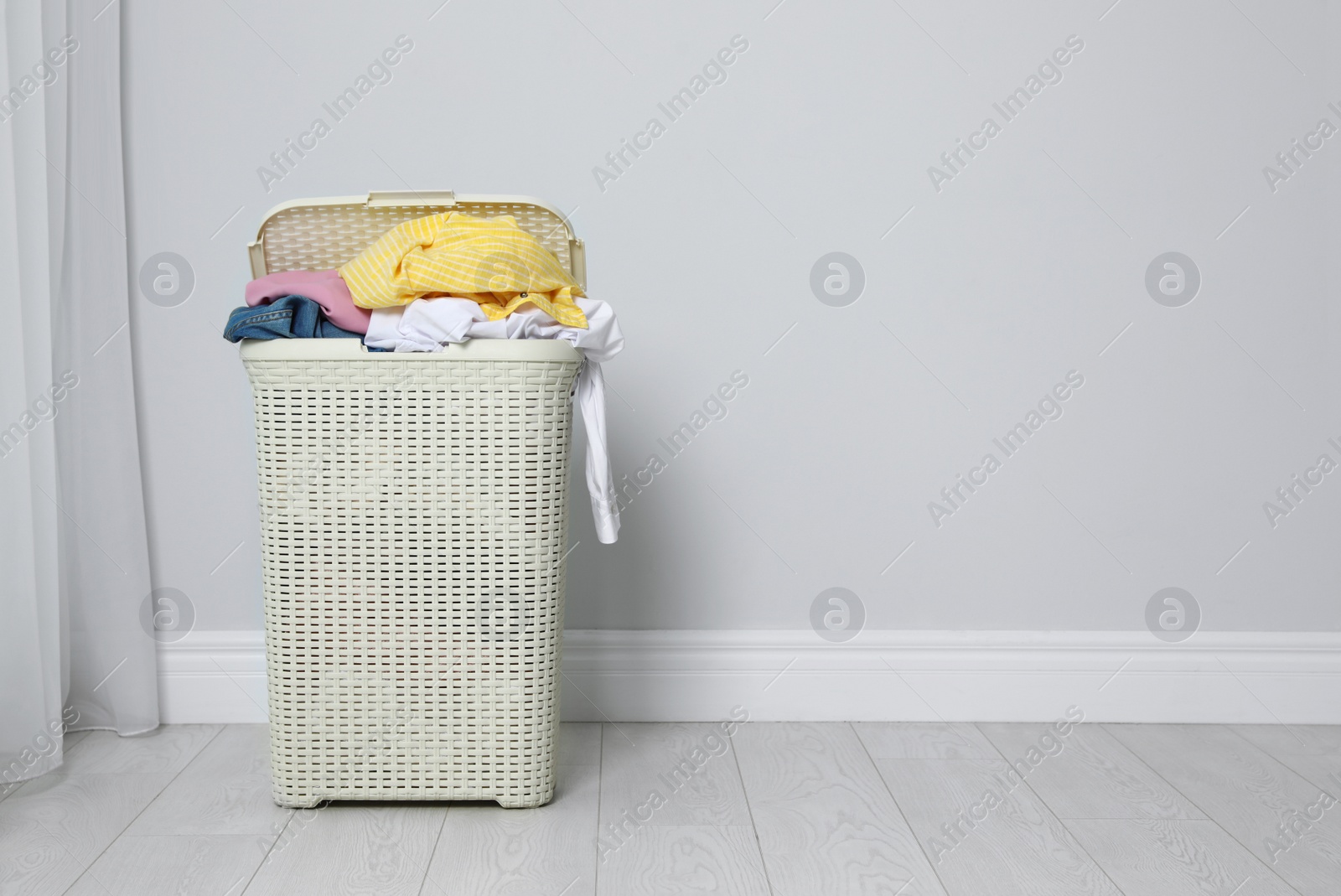 Photo of Plastic laundry basket full of dirty clothes on floor near light wall. Space for text