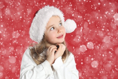 Image of Cute child in Santa hat under snowfall on red background. Christmas celebration
