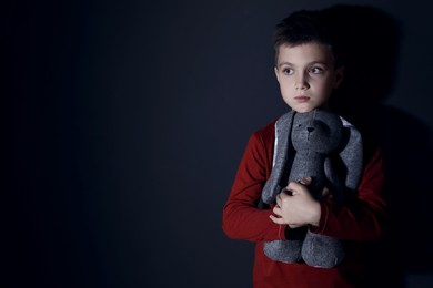 Sad little boy with toy near black wall, space for text. Domestic violence concept