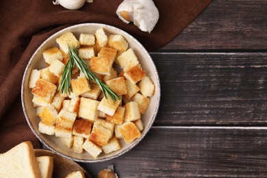 Delicious crispy croutons with rosemary in bowl, toast bread slices and garlic on wooden table, flat lay. Space for text