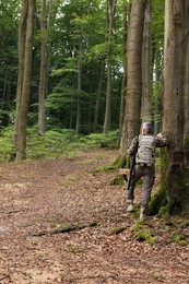 Man with hunting rifle and backpack wearing camouflage in forest, back view. Space for text
