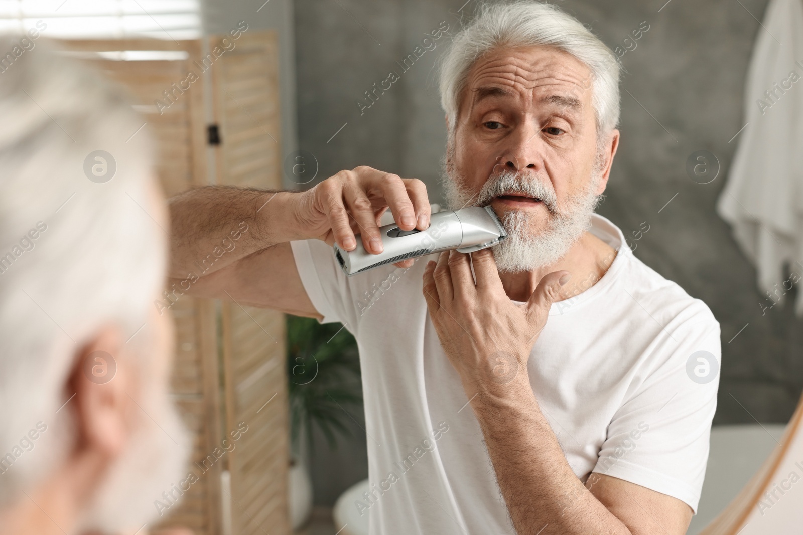 Photo of Senior man trimming beard near mirror in bathroom