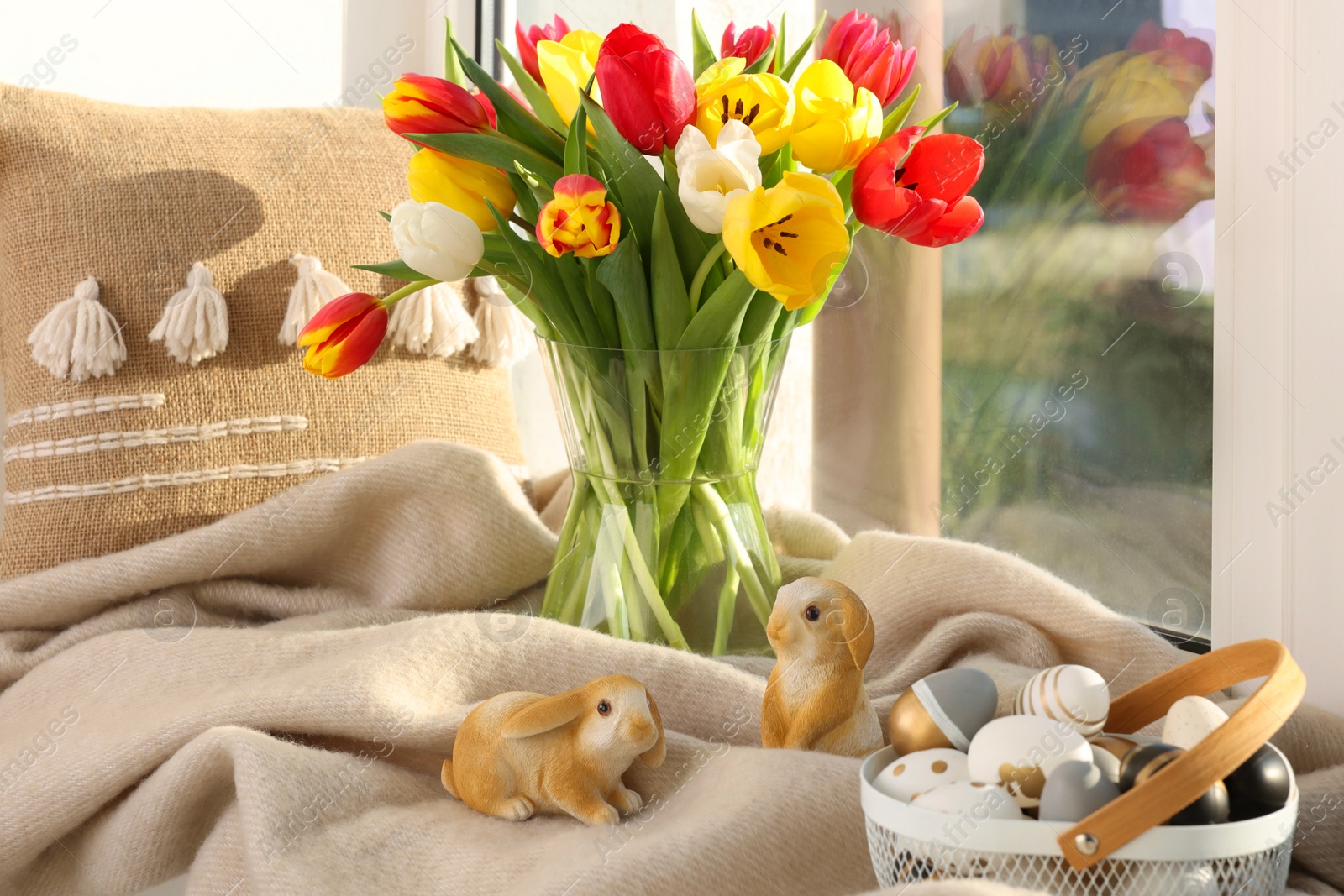 Photo of Easter decorations. Beautiful tulips, basket of painted eggs, bunny figures, pillow and plaid on window sill