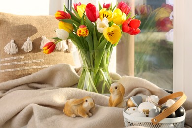 Photo of Easter decorations. Beautiful tulips, basket of painted eggs, bunny figures, pillow and plaid on window sill