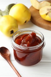 Tasty homemade quince jam in jar, spoon and fruits on white wooden table