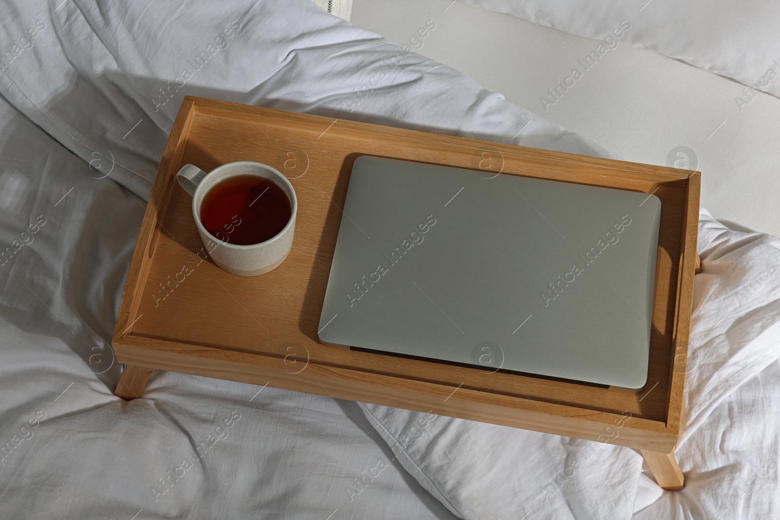 Photo of Wooden tray with modern laptop and cup of aromatic tea on bed indoors, above view