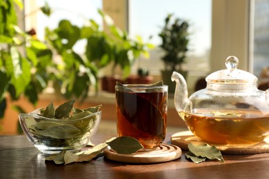 Photo of Cup of freshly brewed tea, bay leaves and teapot on wooden table indoors