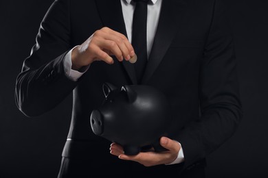 Photo of Businessman putting coin in piggy bank on black background, closeup