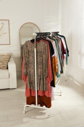 Rack with stylish clothes near sofa and mirror indoors. Fast fashion