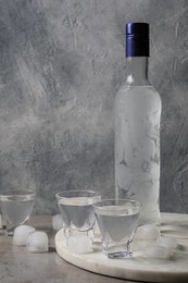 Photo of Bottle of vodka and shot glasses with ice cubes on table against grey wall