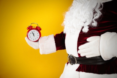 Photo of Santa Claus holding alarm clock on yellow background, closeup. Christmas countdown