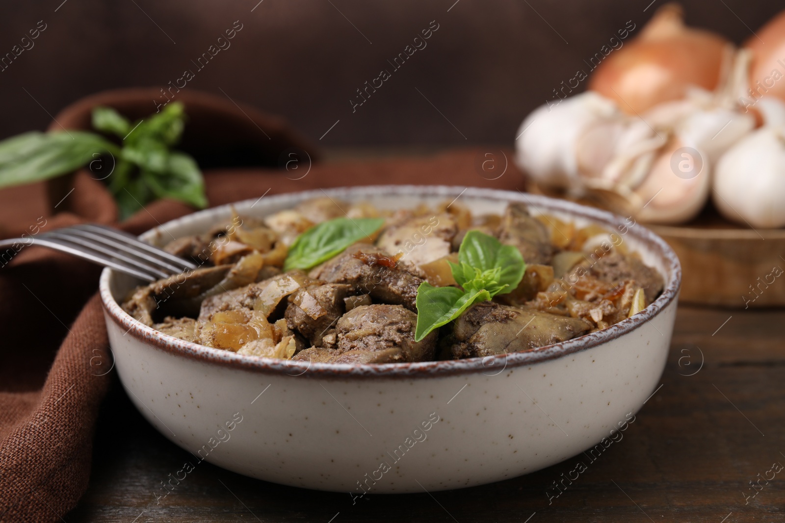 Photo of Delicious fried chicken liver with onion and basil in bowl on wooden table, closeup