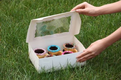 Photo of Woman with box of delicious edible biscuit coffee cups decorated with sprinkles on green grass, closeup