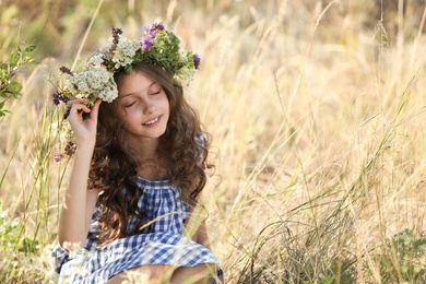 Photo of Cute little girl wearing wreath made of beautiful flowers in field on sunny day