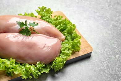 Wooden board with raw chicken breasts and lettuce on grey background